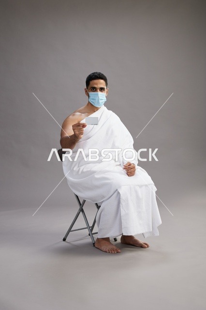 رجل عربي سعودي  خليجي بملابس الإحرام يحمل بطاقة بيضاء فارغه ويرتدي كمامة للوقايه من فايروس كورونا, خلفيه رماديه
