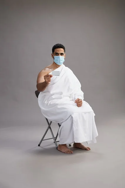 A Saudi Arabian Gulf man in Ihram clothing holding a blank white card and wearing a mask for protection against the coronavirus, with a gray background.