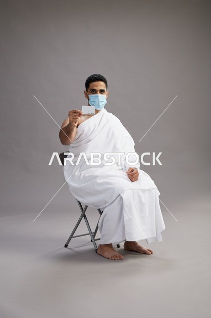رجل عربي سعودي  خليجي بملابس الإحرام يحمل بطاقة بيضاء فارغه ويرتدي كمامة للوقايه من فايروس كورونا, خلفيه رماديه