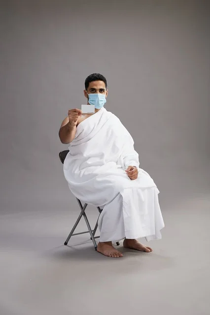 رجل عربي سعودي  خليجي بملابس الإحرام يحمل بطاقة بيضاء فارغه ويرتدي كمامة للوقايه من فايروس كورونا, خلفيه رماديه