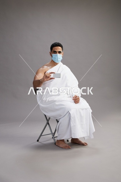 رجل عربي سعودي  خليجي بملابس الإحرام يحمل بطاقة بيضاء فارغه ويرتدي كمامة للوقايه من فايروس كورونا, خلفيه رماديه