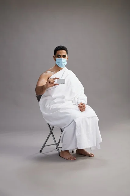 رجل عربي سعودي  خليجي بملابس الإحرام يحمل بطاقة بيضاء فارغه ويرتدي كمامة للوقايه من فايروس كورونا, خلفيه رماديه