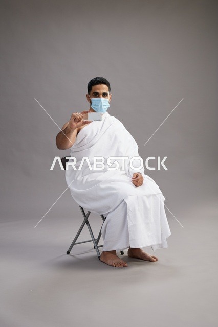 رجل عربي سعودي  خليجي بملابس الإحرام يحمل بطاقة بيضاء فارغه ويرتدي كمامة للوقايه من فايروس كورونا, خلفيه رماديه