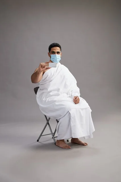 A Saudi Arabian Gulf man in Ihram clothing holding a blank white card and wearing a mask for protection against the coronavirus, with a gray background.