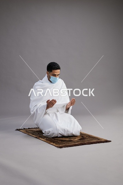 A portrait of an Arab Saudi Gulf man in Ihram clothing wearing a mask, raising his hands in prayer on a prayer rug, performing the rituals of Hajj and Umrah, with a gray background.