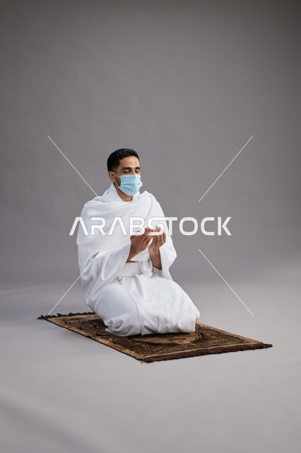 بورتريه لرجل عربي سعودي خليجي بملابس الإحرام يرتدي الكمامه يرفع يديه للدعاء على سجادة الصلاة, اداء مناسك الحج والعمره,  خلفيه رماديه