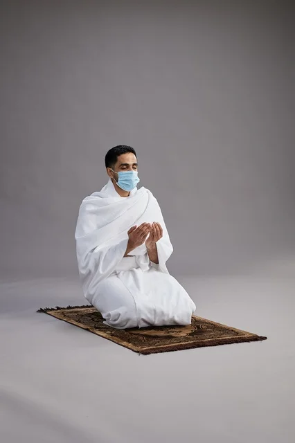 A portrait of an Arab Saudi Gulf man in Ihram clothing wearing a mask, raising his hands in prayer on a prayer rug, performing the rituals of Hajj and Umrah, with a gray background.