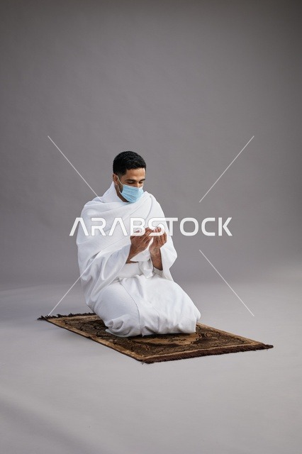بورتريه لرجل عربي سعودي خليجي بملابس الإحرام يرتدي الكمامه يرفع يديه للدعاء على سجادة الصلاة, اداء مناسك الحج والعمره,  خلفيه رماديه