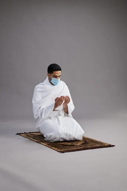 بورتريه لرجل عربي سعودي خليجي بملابس الإحرام يرتدي الكمامه يرفع يديه للدعاء على سجادة الصلاة, اداء مناسك الحج والعمره,  خلفيه رماديه