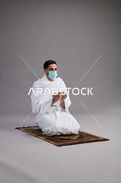 بورتريه لرجل عربي سعودي خليجي بملابس الإحرام يرتدي الكمامه يرفع يديه للدعاء على سجادة الصلاة, اداء مناسك الحج والعمره,  خلفيه رماديه