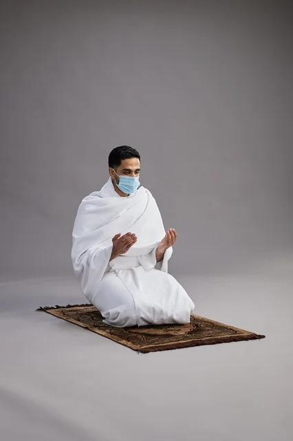 A portrait of an Arab Saudi Gulf man in Ihram clothing wearing a mask, raising his hands in prayer on a prayer rug, performing the rituals of Hajj and Umrah, with a gray background.