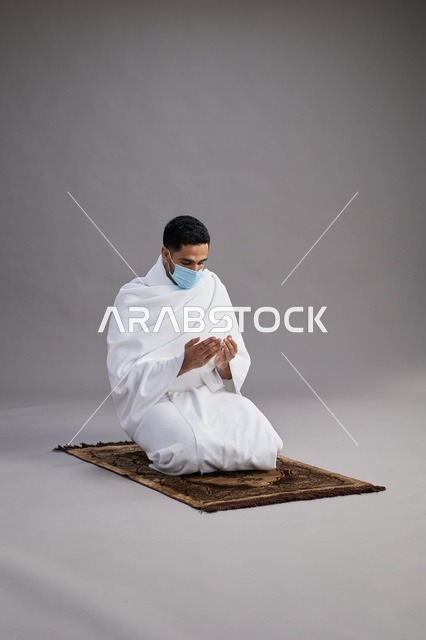 بورتريه لرجل عربي سعودي خليجي بملابس الإحرام يرتدي الكمامه يرفع يديه للدعاء على سجادة الصلاة, اداء مناسك الحج والعمره,  خلفيه رماديه