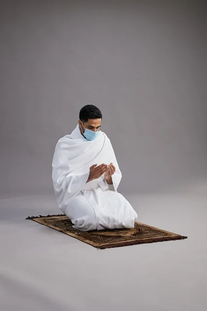 A portrait of an Arab Saudi Gulf man in Ihram clothing wearing a mask, raising his hands in prayer on a prayer rug, performing the rituals of Hajj and Umrah, with a gray background.