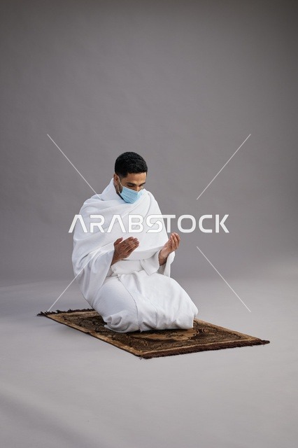 بورتريه لرجل عربي سعودي خليجي بملابس الإحرام يرتدي الكمامه يرفع يديه للدعاء على سجادة الصلاة, اداء مناسك الحج والعمره,  خلفيه رماديه