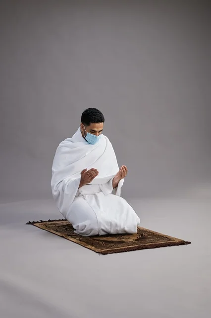بورتريه لرجل عربي سعودي خليجي بملابس الإحرام يرتدي الكمامه يرفع يديه للدعاء على سجادة الصلاة, اداء مناسك الحج والعمره,  خلفيه رماديه