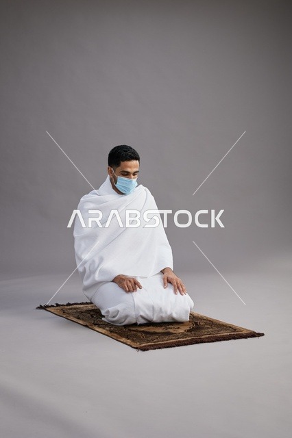 بورتريه لرجل عربي سعودي خليجي بملابس الإحرام يرتدي الكمامه يصلي على سجادة الصلاة, اداء مناسك الحج والعمره,  خلفيه رماديه