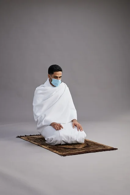 A portrait of an Arab Saudi Gulf man in Ihram clothing wearing a mask, praying on a prayer rug, performing the rituals of Hajj and Umrah, with a gray background.