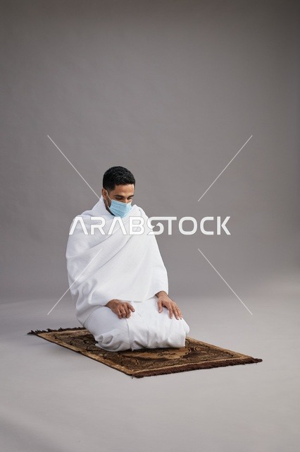 بورتريه لرجل عربي سعودي خليجي بملابس الإحرام يرتدي الكمامه يصلي على سجادة الصلاة, اداء مناسك الحج والعمره,  خلفيه رماديه