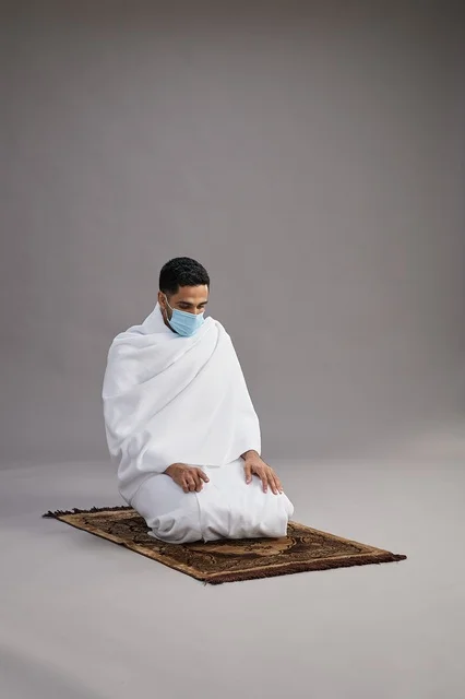 A portrait of an Arab Saudi Gulf man in Ihram clothing wearing a mask, praying on a prayer rug, performing the rituals of Hajj and Umrah, with a gray background.