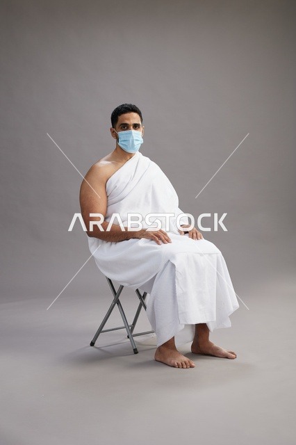 رجل عربي سعودي  خليجي بملابس الإحرام يجلس على كرسي ويرتدي كمامة للوقايه من فايروس كورونا, خلفيه رماديه