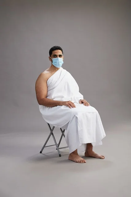A Saudi Arabian Gulf man in Ihram clothing sitting on a chair and wearing a mask for protection against the coronavirus, with a gray background.