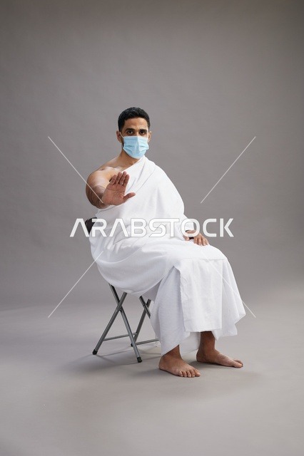 رجل عربي سعودي  خليجي بملابس الإحرام يجلس على كرسي ويرتدي كمامة للوقايه من فايروس كورونا, خلفيه رماديه