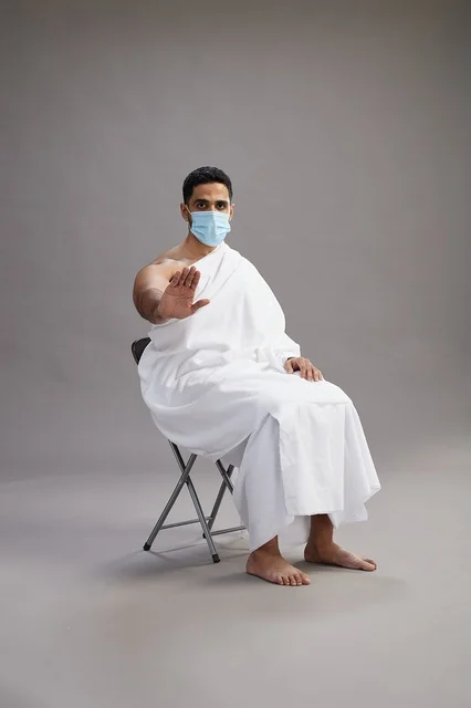 رجل عربي سعودي  خليجي بملابس الإحرام يجلس على كرسي ويرتدي كمامة للوقايه من فايروس كورونا, خلفيه رماديه