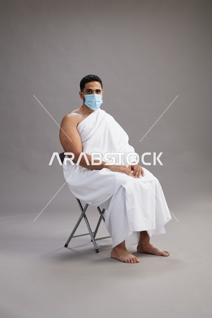 رجل عربي سعودي  خليجي بملابس الإحرام يجلس على كرسي ويرتدي كمامة للوقايه من فايروس كورونا, خلفيه رماديه