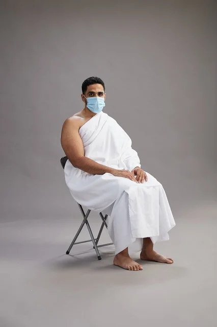 A Saudi Arabian Gulf man in Ihram clothing sitting on a chair and wearing a mask for protection against the coronavirus, with a gray background.