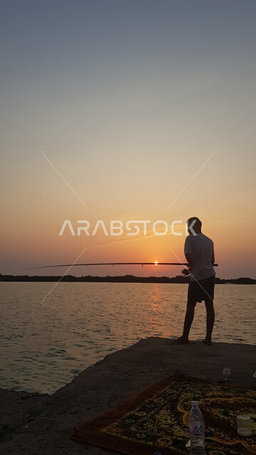 صورة من الخلف لشاب يمارس هواية صيد السمك وقت الغروب على شاطئ كورنيش جدة ، معالم السياحة في المملكة العربية السعودية