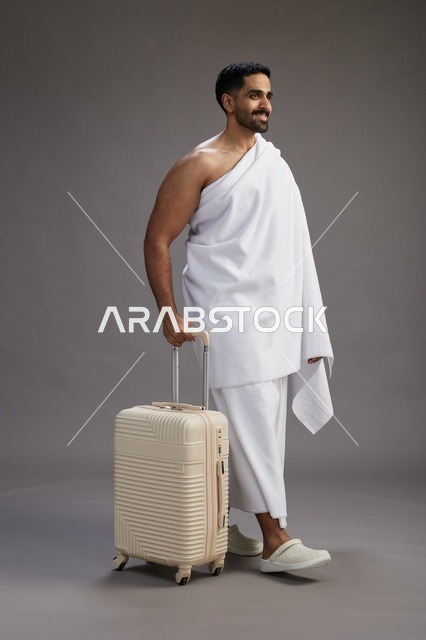 رجل  عربي سعودي خليجي بملابس الإحرام مع حقيبة سفر, الذهاب الى مكه لاداء مناسك الحج والعمره, الجسم بالطول الكامل, خلفيه رماديه