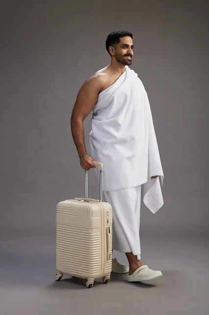 A Saudi Arabian Gulf man in Ihram clothing with a travel bag, going to Mecca to perform the rituals of Hajj and Umrah, full-body, gray background.