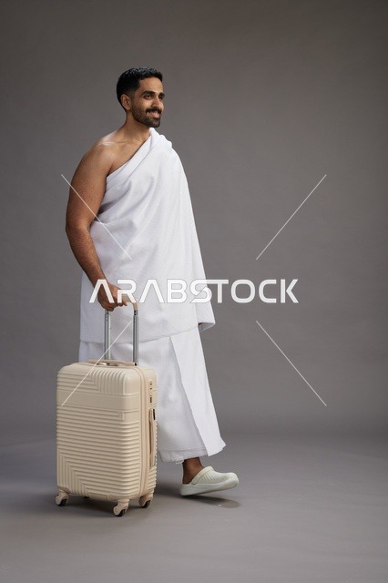 A Saudi Arabian Gulf man in Ihram clothing with a travel bag, going to Mecca to perform the rituals of Hajj and Umrah, full-body, gray background.