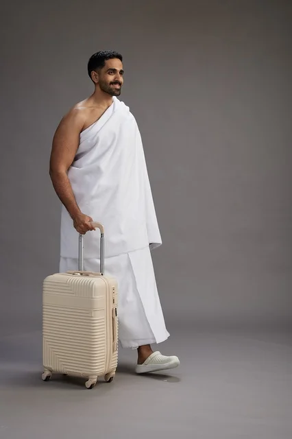 A Saudi Arabian Gulf man in Ihram clothing with a travel bag, going to Mecca to perform the rituals of Hajj and Umrah, full-body, gray background.