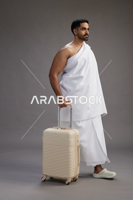 رجل  عربي سعودي خليجي بملابس الإحرام مع حقيبة سفر, الذهاب الى مكه لاداء مناسك الحج والعمره, الجسم بالطول الكامل, خلفيه رماديه