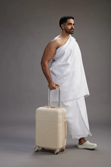 A Saudi Arabian Gulf man in Ihram clothing with a travel bag, going to Mecca to perform the rituals of Hajj and Umrah, full-body, gray background.