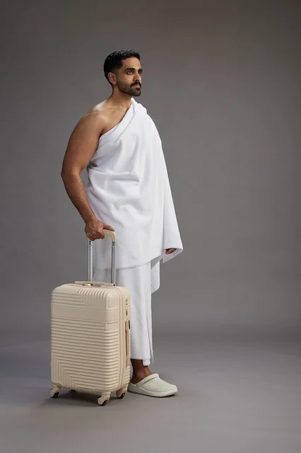 رجل  عربي سعودي خليجي بملابس الإحرام مع حقيبة سفر, الذهاب الى مكه لاداء مناسك الحج والعمره, الجسم بالطول الكامل, خلفيه رماديه