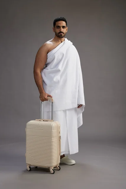 A Saudi Arabian Gulf man in Ihram clothing with a travel bag, going to Mecca to perform the rituals of Hajj and Umrah, full-body, gray background.