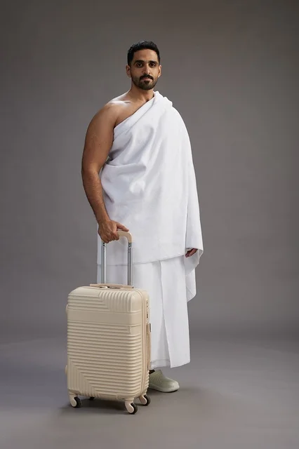 A Saudi Arabian Gulf man in Ihram clothing with a travel bag, going to Mecca to perform the rituals of Hajj and Umrah, full-body, gray background.