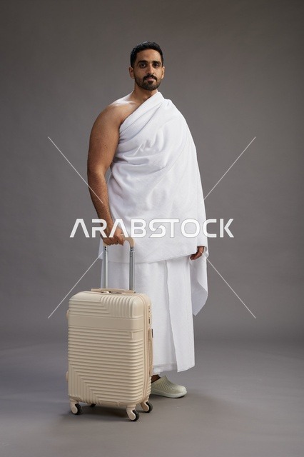 رجل  عربي سعودي خليجي بملابس الإحرام مع حقيبة سفر, الذهاب الى مكه لاداء مناسك الحج والعمره, الجسم بالطول الكامل, خلفيه رماديه
