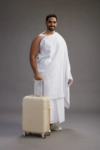 A Saudi Arabian Gulf man in Ihram clothing with a travel bag, going to Mecca to perform the rituals of Hajj and Umrah, full-body, gray background.
