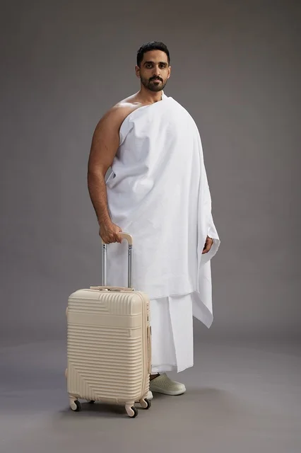 A Saudi Arabian Gulf man in Ihram clothing with a travel bag, going to Mecca to perform the rituals of Hajj and Umrah, full-body, gray background.
