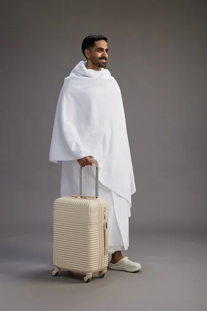 A Saudi Arabian Gulf man in Ihram clothing with a travel bag, going to Mecca to perform the rituals of Hajj and Umrah, full-body, gray background.