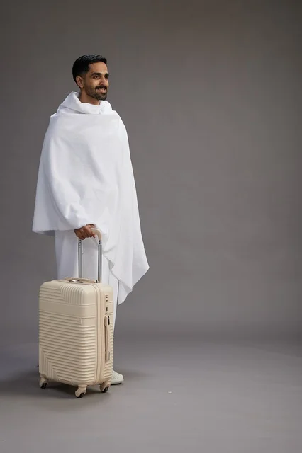 A Saudi Arabian Gulf man in Ihram clothing with a travel bag, going to Mecca to perform the rituals of Hajj and Umrah, full-body, gray background.