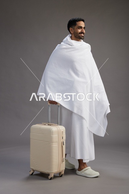 رجل  عربي سعودي خليجي بملابس الإحرام مع حقيبة سفر, الذهاب الى مكه لاداء مناسك الحج والعمره, الجسم بالطول الكامل, خلفيه رماديه