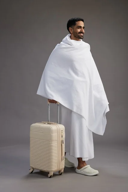 رجل  عربي سعودي خليجي بملابس الإحرام مع حقيبة سفر, الذهاب الى مكه لاداء مناسك الحج والعمره, الجسم بالطول الكامل, خلفيه رماديه