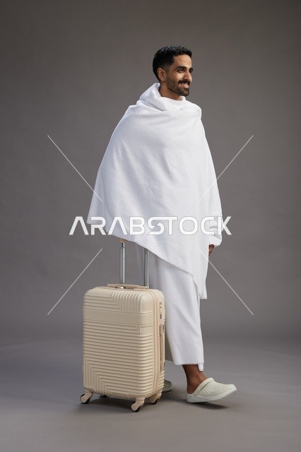 رجل  عربي سعودي خليجي بملابس الإحرام مع حقيبة سفر, الذهاب الى مكه لاداء مناسك الحج والعمره, الجسم بالطول الكامل, خلفيه رماديه