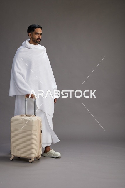 رجل  عربي سعودي خليجي بملابس الإحرام مع حقيبة سفر, الذهاب الى مكه لاداء مناسك الحج والعمره, الجسم بالطول الكامل, خلفيه رماديه