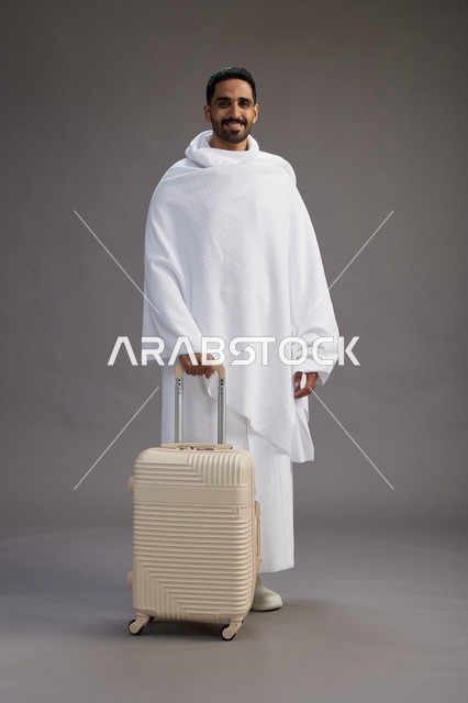 رجل  عربي سعودي خليجي بملابس الإحرام مع حقيبة سفر, الذهاب الى مكه لاداء مناسك الحج والعمره, الجسم بالطول الكامل, خلفيه رماديه