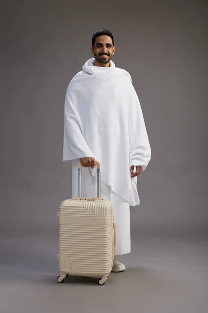 رجل  عربي سعودي خليجي بملابس الإحرام مع حقيبة سفر, الذهاب الى مكه لاداء مناسك الحج والعمره, الجسم بالطول الكامل, خلفيه رماديه